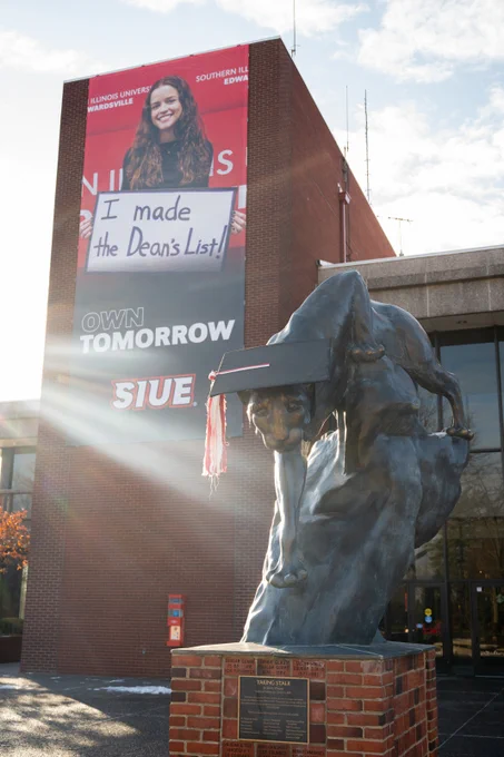 Cougar Statue on a cloudy day wearing a giant mortarboard and tassel. There is a banner outside the MUC framing the statue to the back with a graphic of a graduate holding a sign that reads "I made the Dean's List!"