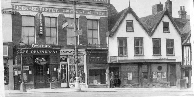 Black and white photo of early 20th-century buildings on a busy street corner, with signs for an oyster bar, restaurant, and various small shops. People stand in doorways and advertisements are visible in shop windows.