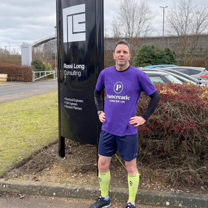 A person in a Pancreatic Cancer UK t-shirt and running clothes is standing next to a sign post with a logo and the words 'Rossi Long Consulting'. Underneath are the words and structural engineers, civil engineers and transport planners
