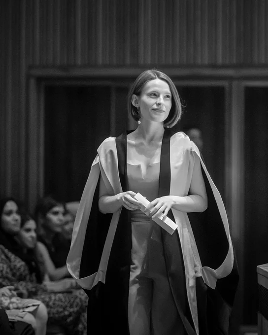 A black and white picture of an alum walking across the stage while holding a graduation scroll.