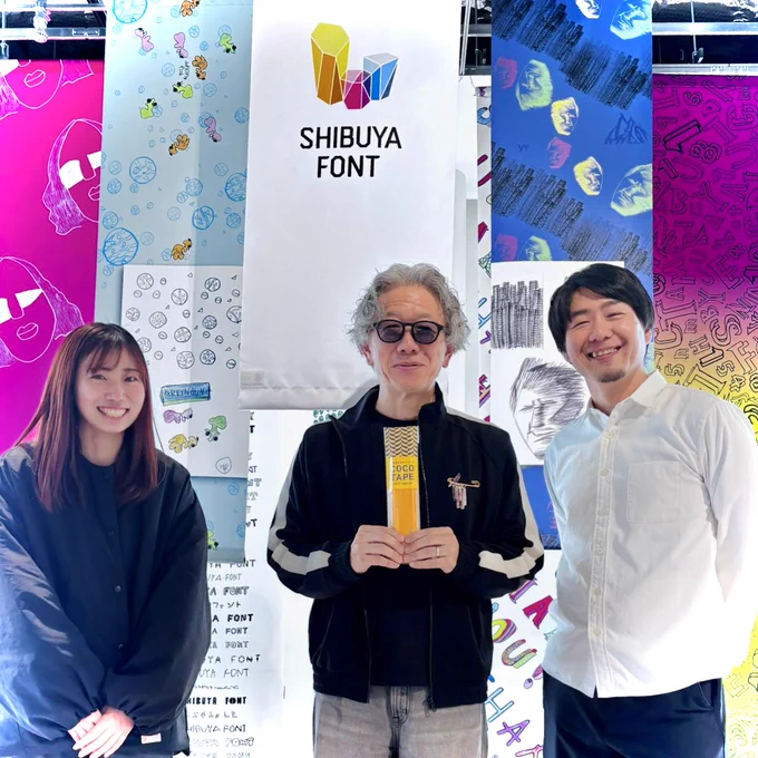 カラフルなシブヤフォントのタペストリーを背景に、シブヤフォントの磯村さん、PLAYWORKSのタキザワ、小島が笑顔で並んでいる。磯村さんは、手に「ココテープ」を持っている。
