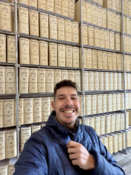 Selfie sonriendo como quien acaba de subir de nivel, con chaqueta azul y gesto de satisfacción, delante de estanterías repletas de enormes libros de protocolo notarial perfectamente ordenados, en una sala que huele a papeleo serio y a decisiones importantes.