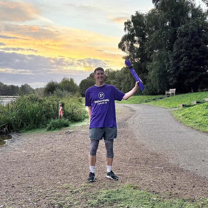 A person standing next to Whitlingham Broad in a Pancreatic Cancer UK t-shirt, holding a Pancreatic Cancer UK bam bam stick in the air. The sun is rising behind them.
