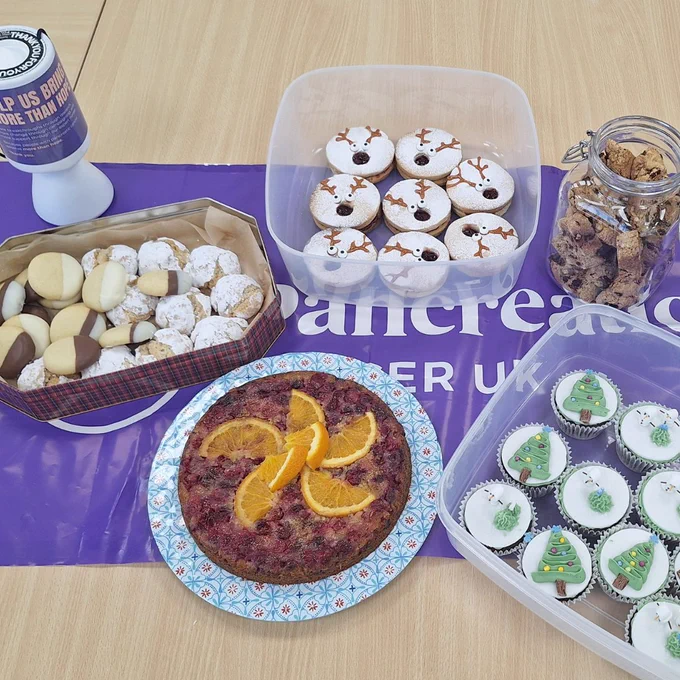 A table of Christmas themed cakes and biscuits on a Pancreatic Cancer UK table cloth, with a Pancreatic Cancer UK donation pot.