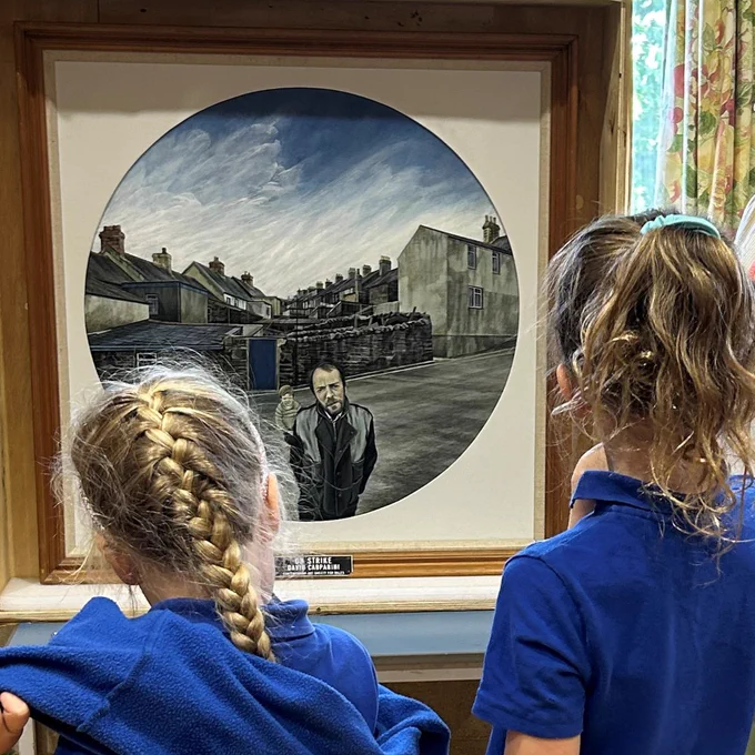 Two children in blue school uniforms look at a framed circular painting. The painting shows a man and a little boy standing on a street with rows of terraced houses behind them.
