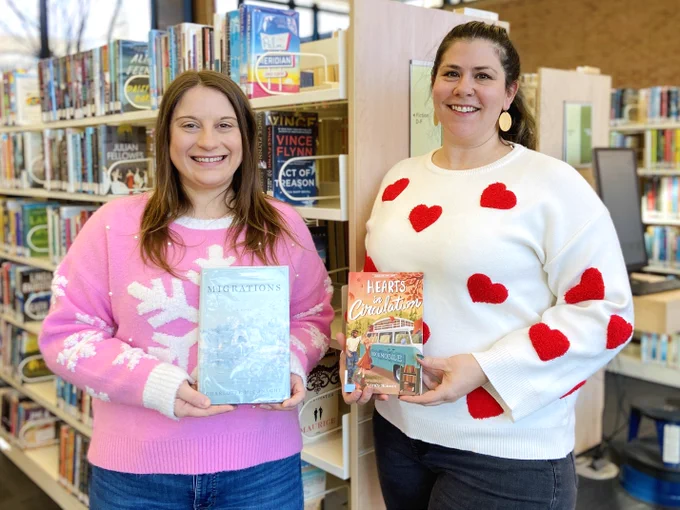Two staff members pose with books; one in a pink snowflake sweater and another wearing red hearts.