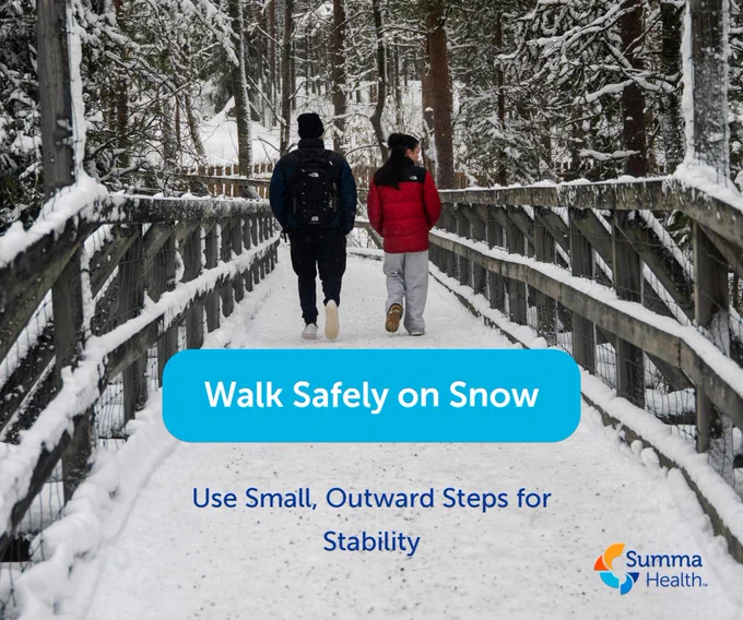 Two people walk carefully on a snow-covered wooden bridge surrounded by snow-laden trees in winter.