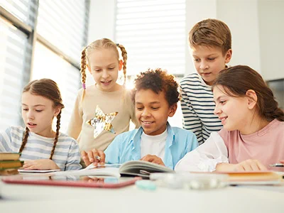 Group of five children gathered around a table, engaged in reading and learning activities in a bright classroom.