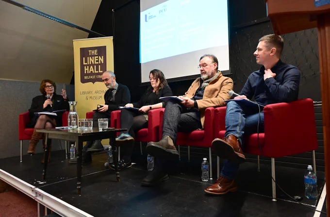 Five panelists seated on red chairs on a stage at The Linen Hall Library, engaged in a discussion with a presentation screen behind them.