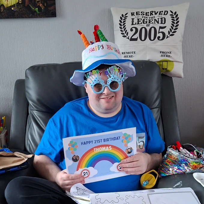 Man wearing a colourful birthday hat and blue shirt holding a 21st birthday card with a rainbow and balloons design.