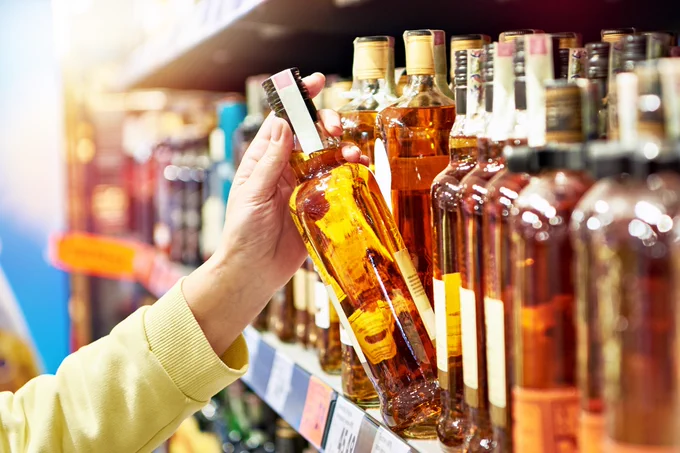 A person examining a bottle of liquor in a store, with a variety of other bottles on the shelves in the background.