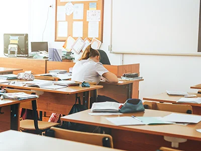 Student sitting alone at a desk in a quiet classroom filled with scattered papers and books.