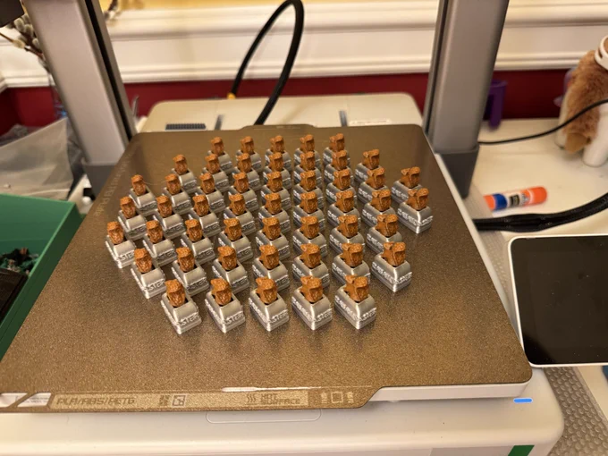 Alt text:
Dozens of small, identical 3D-printed parts arranged in a neat grid on a gold textured 3D printer build plate, each with a silver toaster shaped base and a brown, squirrel on top, inside a desktop 3D printer. Caption by ChatGPT