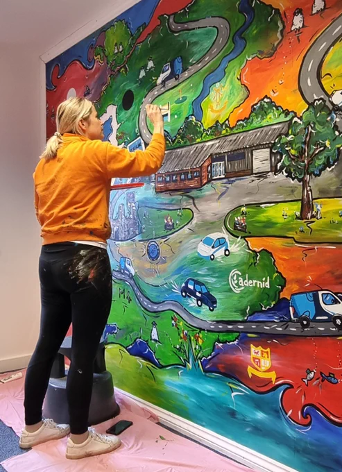A local artist updating the painted vibrant wall mural originally commissioned a decade earlier inside BCC IT's office in Newcastle Emlyn, Wales. It depicts a stylised local landscape of BCC IT, winding roads, the river Teifi, castle ruins, The river Severn and Severn bridge, and surrounding countryside.