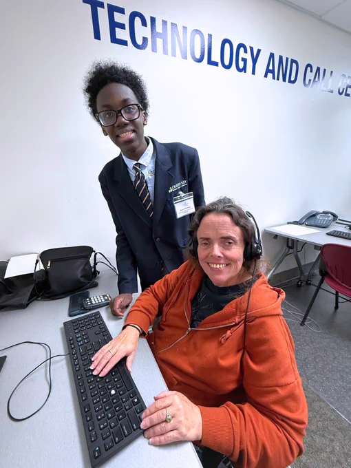 A photo of two people smiling for the camera at Miami Lighthouse for the Blind in the computer lab: an adult vocational rehabilitation participant from Miami Lighthouse for the Blind sits as student volunteer Omari stands nearby, teaching them how to use the computer with visual impairment.