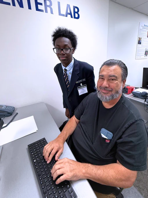 A photo of two people smiling for the camera at Miami Lighthouse for the Blind in the computer lab: an adult vocational rehabilitation participant from Miami Lighthouse for the Blind sits as student volunteer Omari stands nearby, teaching them how to use the computer with visual impairment.