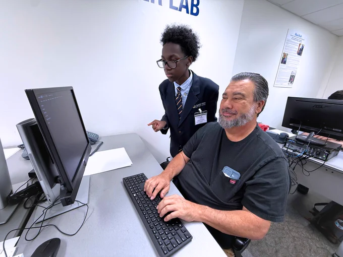 A photo of two people posing for the camera at Miami Lighthouse for the Blind in the computer lab: an adult vocational rehabilitation participant from Miami Lighthouse for the Blind sits at a computer with their hands on the keyboard as student volunteer Omari stands nearby, teaching them how to use the computer.