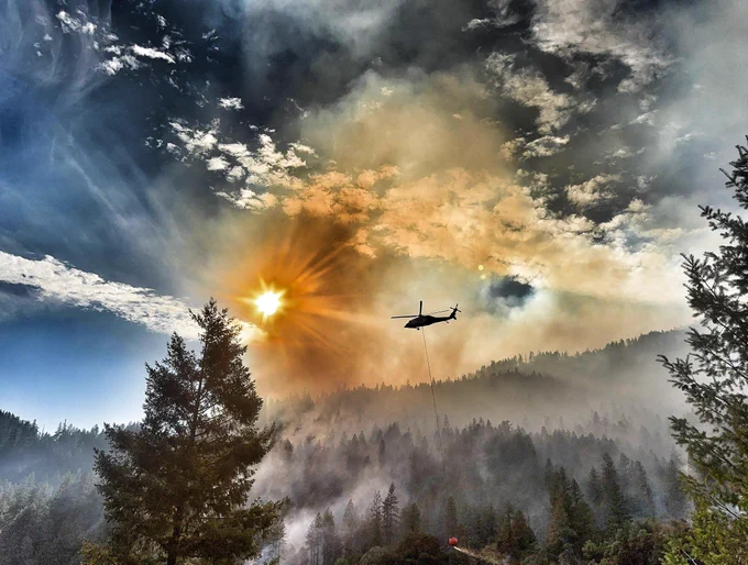 Helicopter flying over a wildfire with smoke billowing under a bright sun in a cloudy sky. Photo by Jodi Plotcher. 