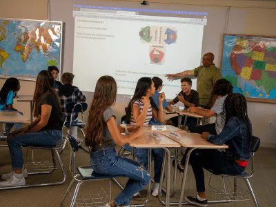 A classroom scene with students sitting at tables and a teacher standing near a whiteboard displaying a website with educational logos. Some students are engaged in discussion while others are using laptops. A world map is visible in the background.