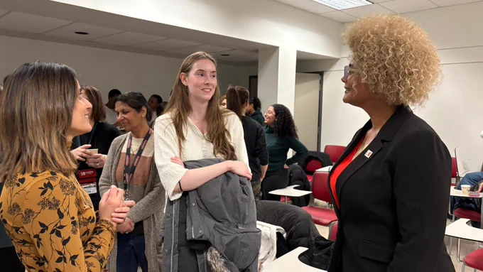 Two women engaged in a conversation with Dean Andrade, with other attendees in the background.