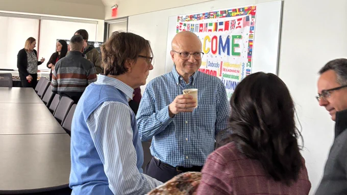 Group of faculty members, one holding a coffee, mingling in a room with a "Welcome" banner on the wall, with other faculty and staff in the background.