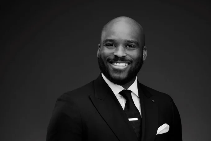 A black and white image of a person, smiling and wearing a dark suit. 
