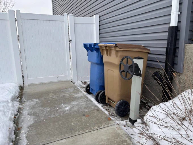 Two wheeled garbage bins stand next to a house. 