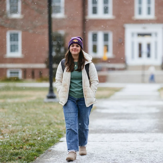 A person wearing a white jacket, jeans, and a purple beanie walks on a snowy path with a brick building in the background.