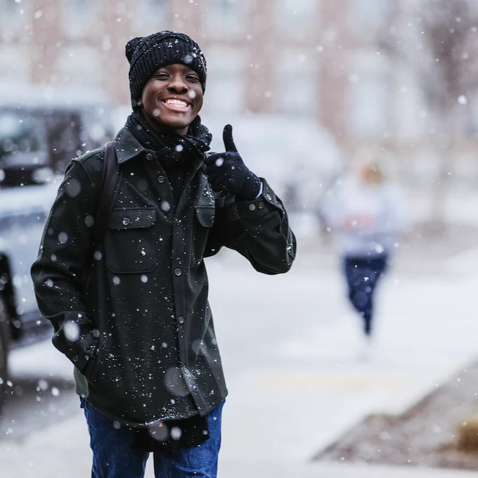 Person in a winter coat and beanie smiling and giving a thumbs up in a snowy setting.