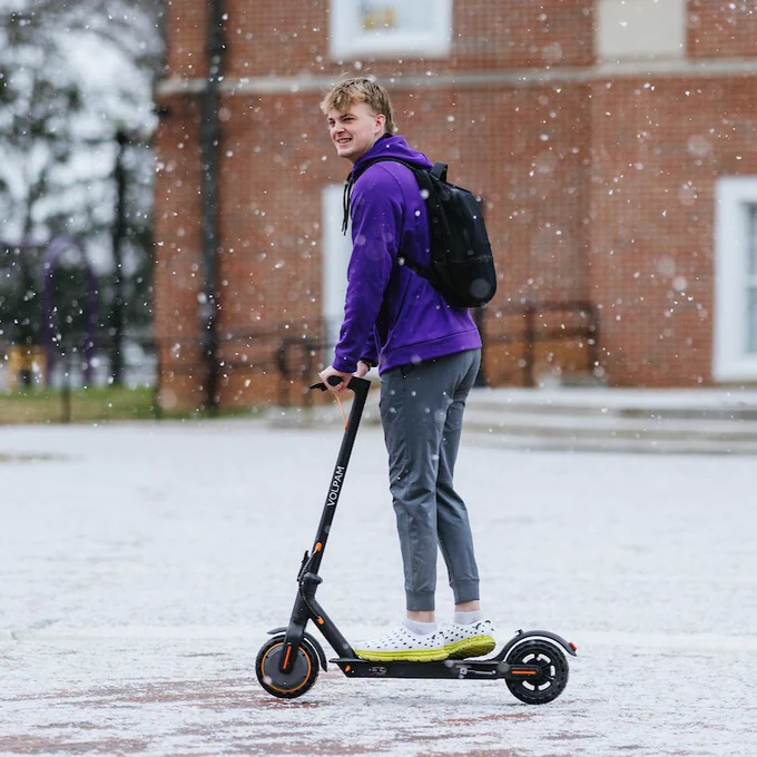 A person in a purple hoodie and gray pants stands with a scooter on a snowy day, smiling slightly towards the camera.
