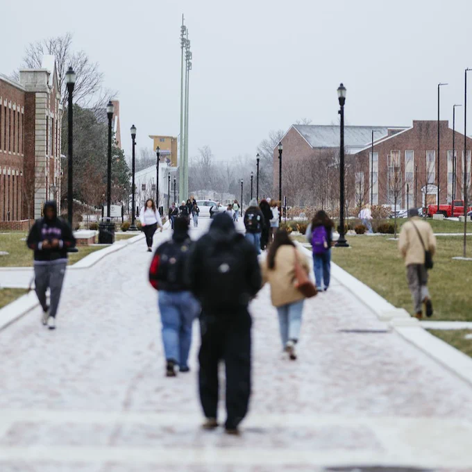 Several people walking on a university campus pathway, with buildings on both sides and a cloudy sky overhead.