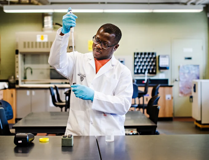 Augustine Osei working in a lab at the University of Waterloo. Soil scientists work in the outdoors, labs, and offices. Credit: Hilary Gauld, Ecology lab in the Faculty of Environment, University of Waterloo.  Image description: a man wearing a lab coat and blue gloves stands a chemistry bench holding a pipette 