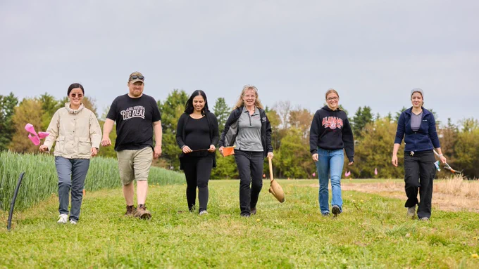 Six people walk across a grassy research field holding flags and sampling tools, with crop plots and trees in the background.