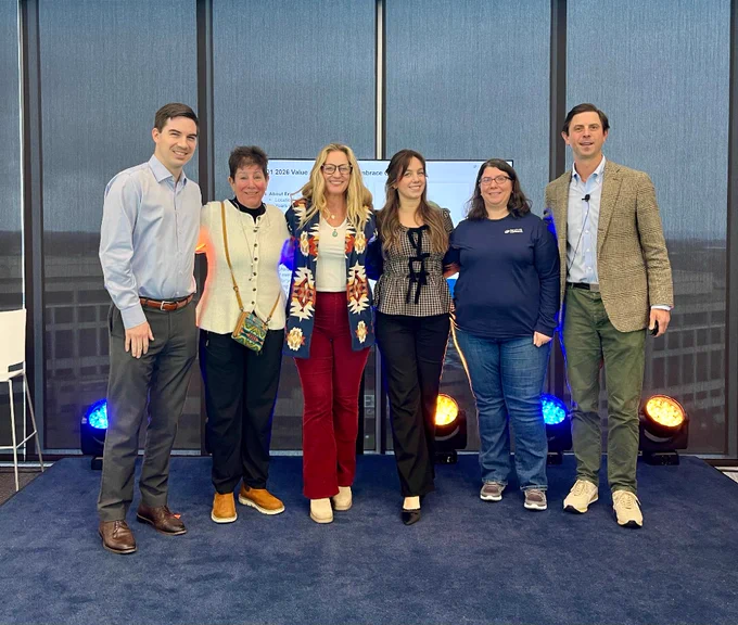 A group photo taken on stage at ConstructConnect’s Q1 Town Hall, showing five Values Award winners standing between two company leaders. They are smiling in front of tall windows with a city view in the background. Stage lights in blue and orange shine behind them, adding a celebratory feel. The group is dressed in business-casual clothing, standing close together to mark the moment of recognition for living the company values of People First, Deliver Customer Value, Expand Your Impact, Make It Happen, and Embrace Change.


