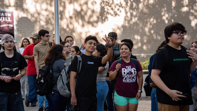 Two TAMIU students acknowledging the camera and smiling while standing in line with other students.