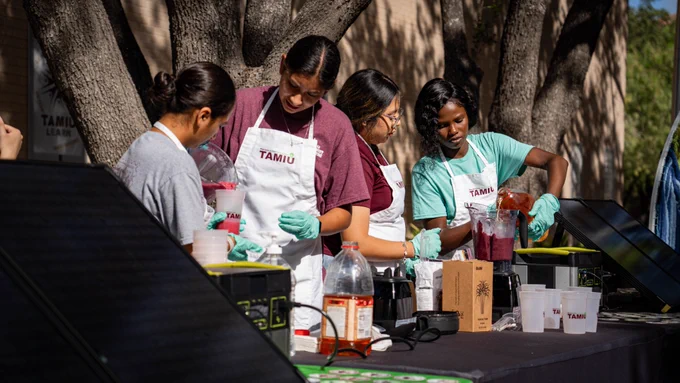 Four TAMIU students wearing aprons that say "TAMIU" making smoothies with solar panels on the table.