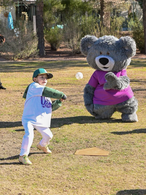 A child hitting a baseball. Cuddles is acting as the catcher behind home plate. They are in an outdoor setting.