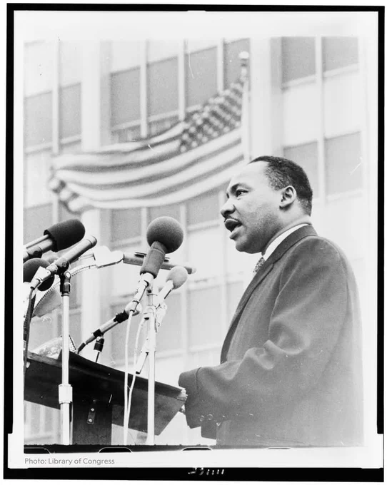 Martin Luther King Jr. speaking into multiple microphones at a podium with an American flag in the background.