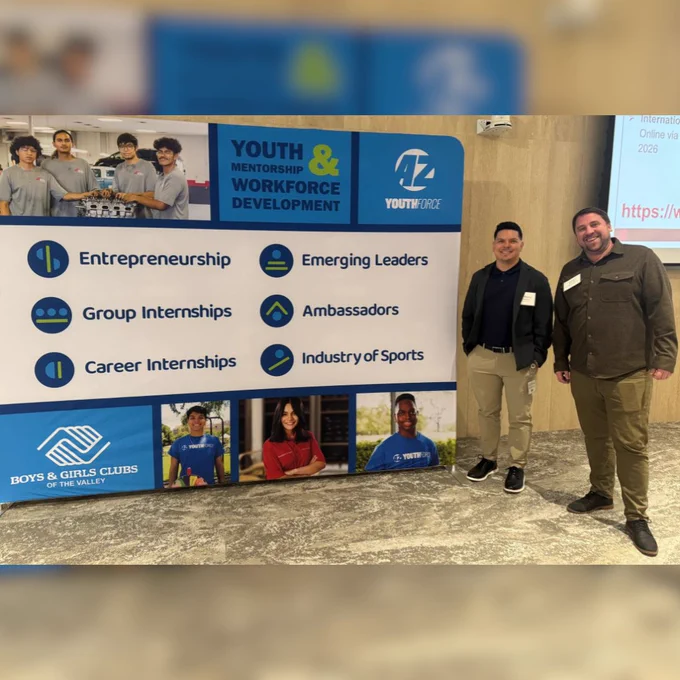 Anthony Amezquita and Julian Cashman stand next to a large blue and white display for the Boys & Girls Clubs of the Valley AZ YouthForce program. The backdrop highlights "Youth Mentorship & Workforce Development" and lists key program areas such as Entrepreneurship, Group Internships, and Career Internships. Anthony is wearing a navy polo with a grey blazer and khakis, while Julian is wearing a dark olive button-down shirt and olive trousers.