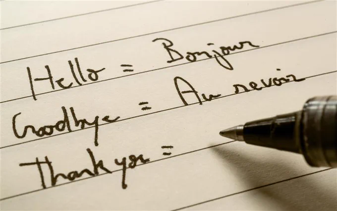 A black ink pen lifted slightly from yellow lined paper. On the paper are handwritten notes which read, "Hello = Bonjour Goodbye = Au Revoir Thank you =" 