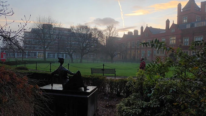 A quadrangle with lawns and trees in the middle-ground and a mix of modern and Victorian buildings in the background, viewed from behind a statue and garden bushes.
