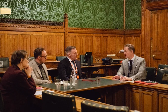 Four people sit around a table in a wood-panelled parliamentary room, in discussion during a meeting.