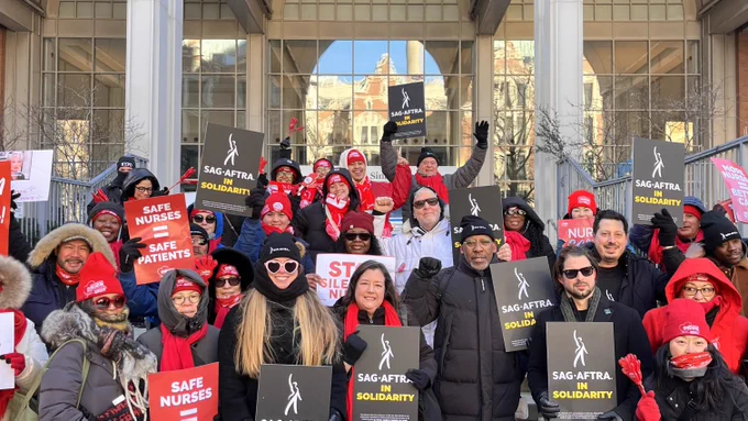 A group of people wearing mostly red, white and black stand on the stairs of a building. They hold various signs that read "Safe nurses = safe patients," "Nurses Deserve Good Health Benefits," and "Sag-Aftra in Solidarity."