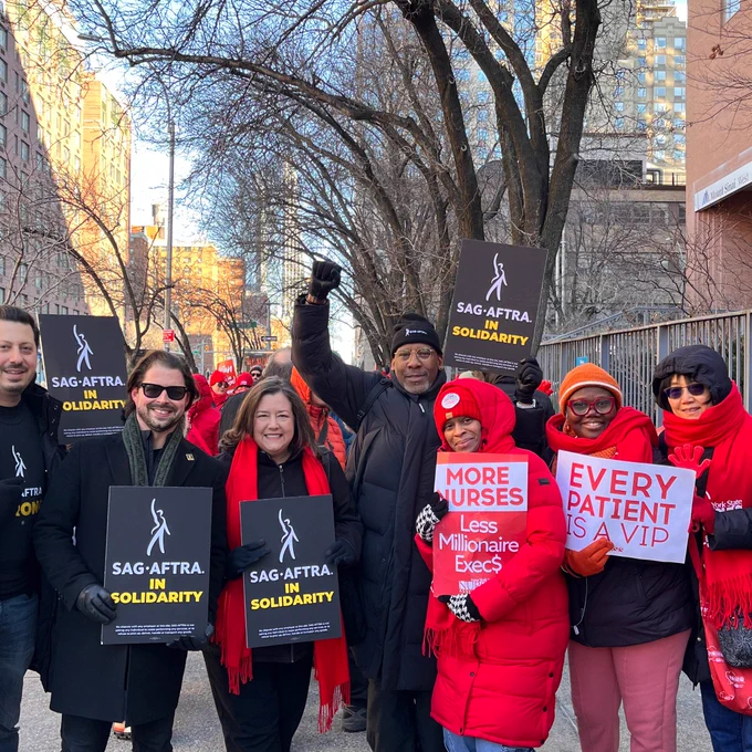 A group of people wearing black and red stand in a line. They hold signs that read "Sag-Aftra in Solidarity," "More Nurses, less millionaire execs," and "Every Patient is a VIP."