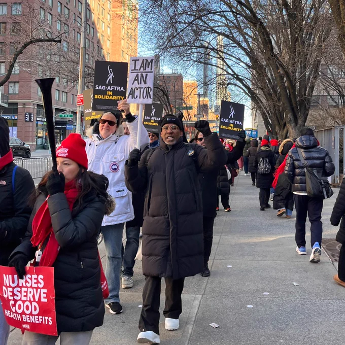 A group of people walk on a sidewalk searing black, red, and white. They hold various signs that read, "Nurses deserve good health behefits," "Sag-Aftra in Solidarity," and "I Am An Actor."
