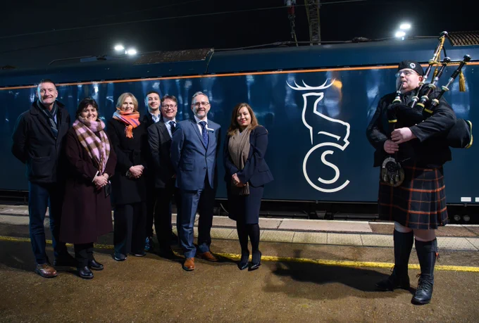 Group of formally dressed people standing on a train platform at night next to the locomotive.