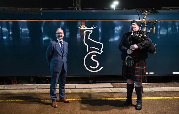 Man in traditional Scottish attire playing bagpipes next to a suited man standing on a train platform at night.