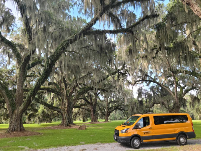 Yellow USGS field van parked on green grass beneath large oak trees draped with Spanish moss on a sunny day.