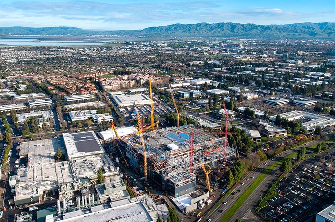 Wide helicopter aerial of a large construction site in Sunnyvale, California, with multiple cranes and a steel-framed building; city and mountains in the background.