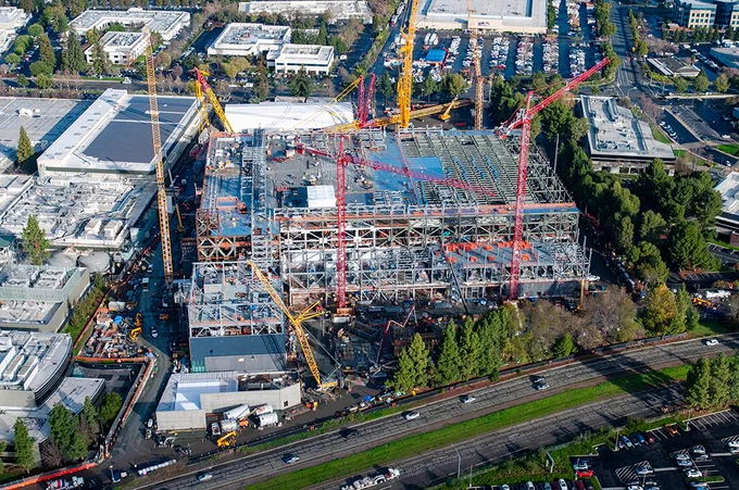 Closer helicopter aerial of the same construction site showing the steel structure, cranes, and active work areas around the building footprint.